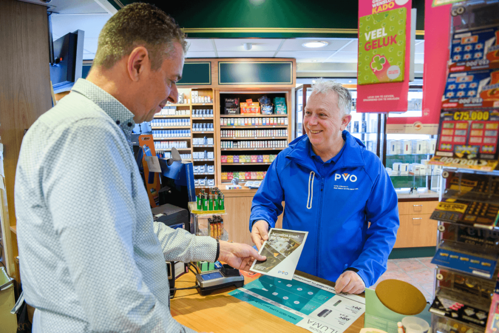 Medewerker van PVO Zeeland West-Brabant met een blauwe PVO-jas staat aan de kassa in een tabakszaak. De PVO medewerker overhandigt een folder van PVO aan de man achter de kassa.