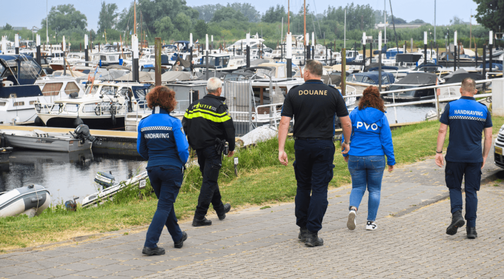 Medewerkers van de handhaving, politie, PVO Zeeland West-Brabant en de Douane lopen langs een haven waarin boten liggen.