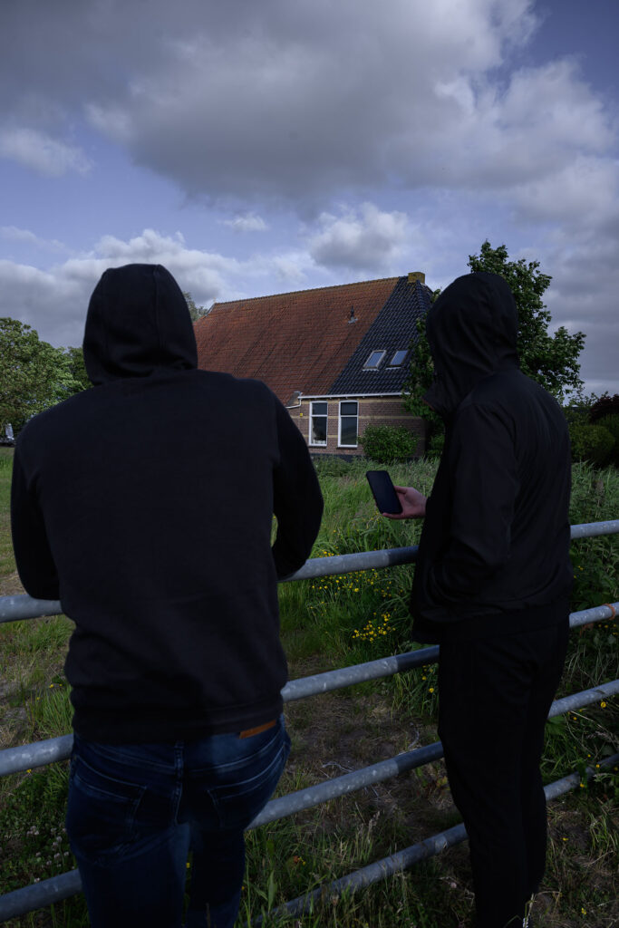 Twee personen met donkere kleding die naar een boerderij kijken. De lucht is donker en er hangt een dreigende sfeer.