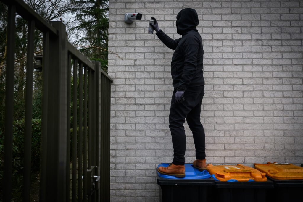 Persoon in donkere kleding met een capuchon op die op een kliko staat om een beveiligingscamera te bespuiten met een spuitbus verf.